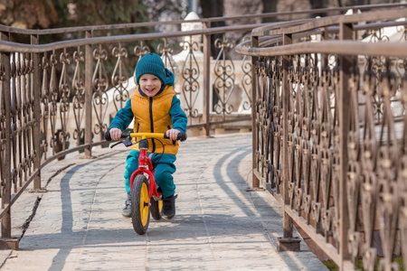 A cheerful little boy rides a bicycle outdoors. A happy child walks in the spring park. The baby is dressed in a fashionable yellow vest and turquoise jumpsuit.の写真素材