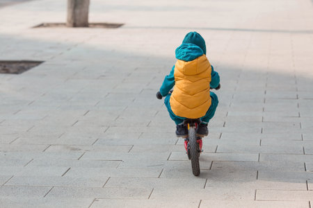 A cheerful little boy rides a bicycle outdoors. A happy child walks in the spring park. The baby is dressed in a fashionable yellow vest and turquoise jumpsuit.の写真素材