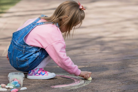 A happy girl in pink clothes draws a rainbow with chalk on the asphalt on the street. Portrait of a...