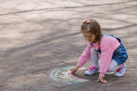 A happy girl in pink clothes draws a rainbow with chalk on the asphalt on the street. Portrait of a little girl drawing with rainbow chalk on a sunny summer day. creative development of children.の写真素材