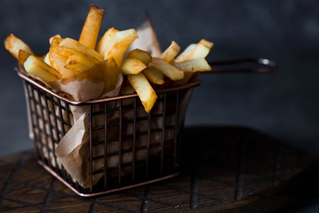 French fries in a metal basket with salt on a stylish board on a dark background. Fried potatoes. The concept of fast food and junk food.の写真素材