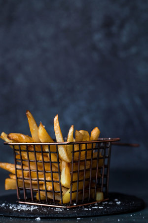 French fries in a metal basket with salt on a stylish board on a dark background. Fried potatoes. The concept of fast food and junk food.の写真素材