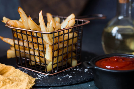 French fries in a metal basket with salt, ketchup and sunflower oil on a stylish board on a dark background. Fried potatoes. The concept of fast food and junk food.の写真素材