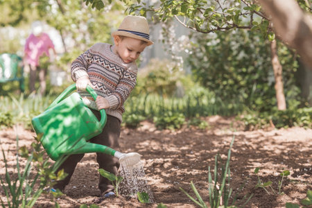 A cute boy in a hat plants and waters seedlings from a watering can in a summer garden, outdoors. The concept of gardening and teaching a child to work.の写真素材