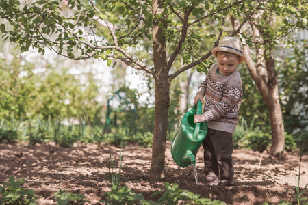 A cute boy in a hat plants and waters seedlings from a watering can in a summer garden, outdoors. The concept of gardening and teaching a child to work.の写真素材