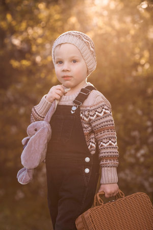 A little boy of 4 years old outdoors with a suitcase and a stuffed rabbit goes on a journey. A beautiful portrait of a child playing on the street.の写真素材