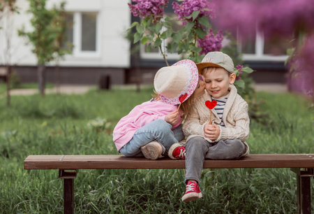 Lifestyle. Little beautiful children on a bench with candy lollipops on a stick in the shape of a heart. A girl kisses a boy. The concept of childhood friendship and first love.の写真素材
