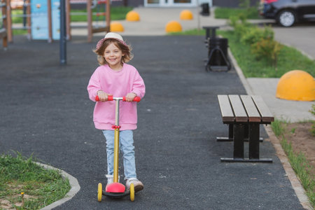A cheerful little beautiful girl rides a scooter outdoors on the playground. A happy child walks in the fresh air. The baby is dressed in a fashionable pink hoodie and jeans.の写真素材