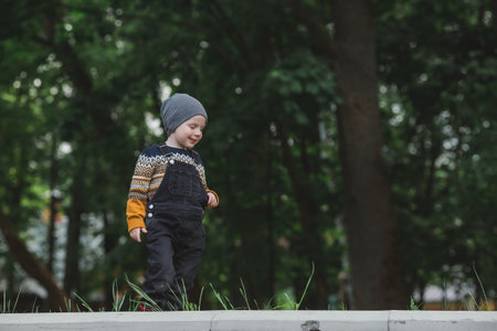 Portrait of a fashionable little boy outdoors. A happy child walks in the spring park. The kid is dressed in a fashionable sweater, sneakers and denim overalls.の写真素材