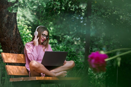 A young, happy, beautiful freelance businesswoman sitting on a park bench with headphones and a laptop. A woman works outdoors or makes purchases in an online store.の写真素材