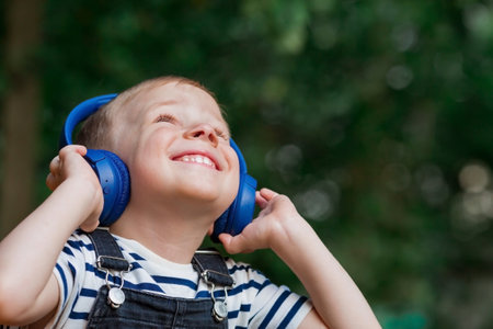 Cute, cheerful, little boy smiles and listens to music in big blue headphones in nature in the park. Music for children's development.の写真素材