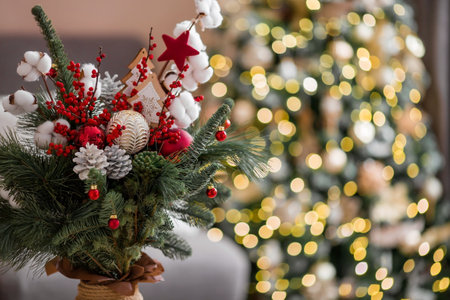 A bouquet of fir trees with red berries, cotton, toys on the table against the background of Christmas tree garlands. Winter decorations in the interior. The concept of preparation for the New Year holidays 2024.の写真素材