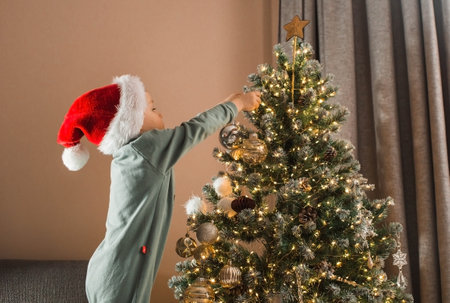 A beautiful little boy of 4 years old in a Santa Claus hat is decorating a Christmas tree in the living room. Christmas Eve. The concept of celebrating Christmas and New Year 2024.の写真素材