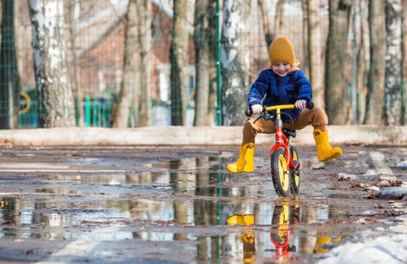 A cheerful little boy rides a bike through puddles. A happy child walks outside in the spring. The kid is dressed in a fashionable plaid shirt, overalls and yellow rubber boots.の写真素材