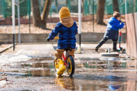 A cheerful little boy rides a bike through puddles. A happy child walks outside in the spring. The kid is dressed in a fashionable plaid shirt, overalls and yellow rubber boots.の写真素材