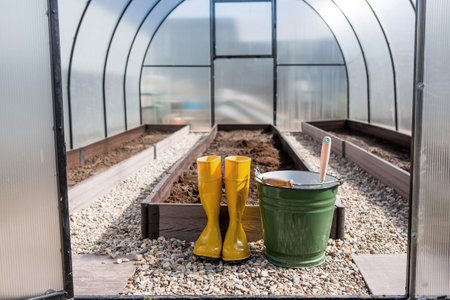 Yellow rubber boots, a bucket and a small shovel in the new greenhouse. The concept of spring planting of vegetables and farming.の写真素材