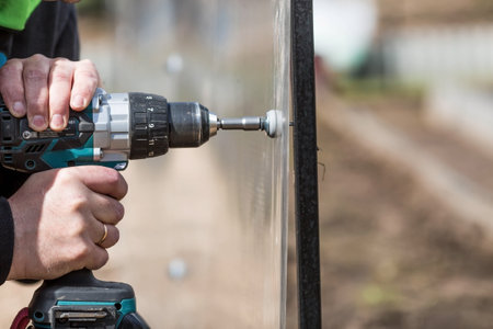 A man holds a battery-powered screwdriver and assembles a new greenhouse made of polyethylene, polycarbonate. Macro photography. Side view Preparing for the spring garden season.の写真素材