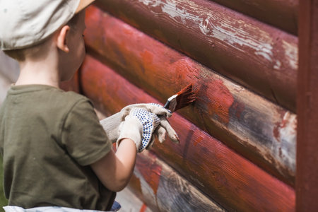 A little boy is working with a brush and applying red paint to a wooden village house. Teaching a child to work. Half of the painted surface. Smearing with a brush.の写真素材