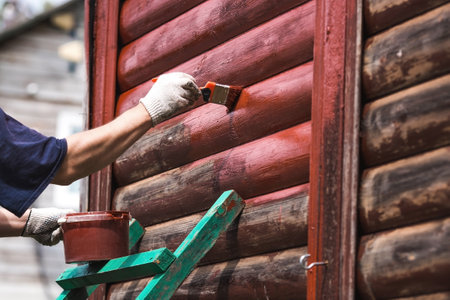 A man works with a brush and applies red paint to a wooden village house. A worker paints wooden planks in close-up. Half of the painted surface. Smearing with a brush.の写真素材