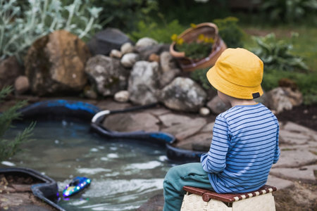 A 5-year-old boy in a yellow panama hat launches a radio-controlled boat in a pond. Children's fun and street games. Child's leisure time.の写真素材
