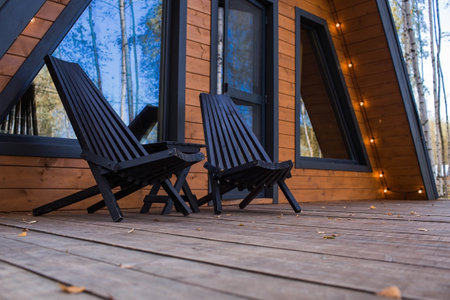 Two stylish wooden deck chairs on the veranda of an A-shaped frame house in Scandinavian style. Quiet rest. The concept of glamping and chalet rentals.の写真素材