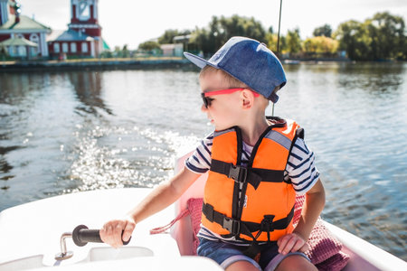 A handsome, fashionable little boy of 5 years old in a bright life jacket and sunglasses drives a catamaran on the lake. Recreation and tourism.の写真素材