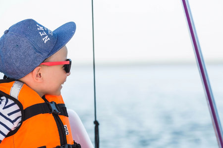 A handsome, fashionable little boy of 5 years old in a bright life jacket and sunglasses drives a catamaran on the lake. Recreation and tourism.の写真素材