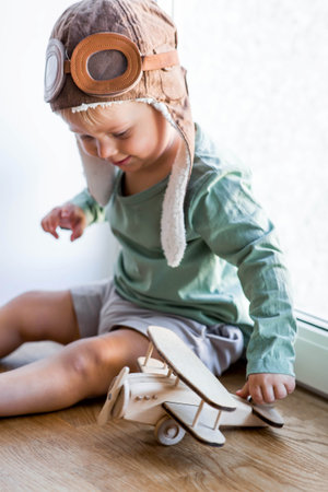 A cute 2-year-old boy in a cap is playing on the windowsill with a wooden airplane. Children's...