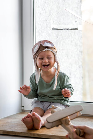 A cute 2-year-old boy in a cap is playing on the windowsill with a wooden airplane. Children's...