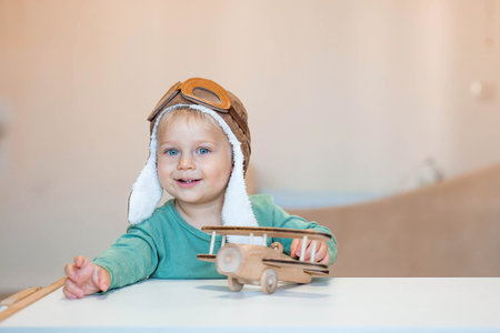 A cute 2-year-old boy in an aviator pilot's hat is playing with a wooden airplane. Children's wooden toys for child development.の写真素材