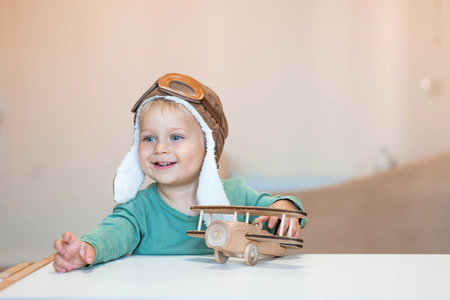 A cute 2-year-old boy in an aviator pilot's hat is playing with a wooden airplane. Children's wooden toys for child development.の写真素材