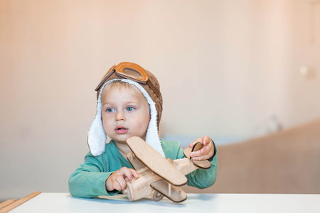 A cute 2-year-old boy in an aviator pilot's hat is playing with a wooden airplane. Children's wooden toys for child development.の写真素材