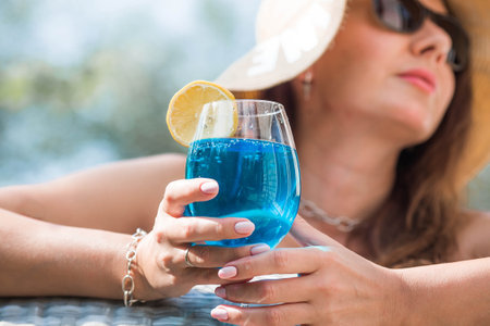 A young woman in a swimsuit and sunglasses in a bathtub with a glass of Blue Lagoon cocktail on the background of the pool. The concept of a summer vacation, vacation and foam party.の写真素材