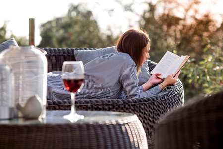 A woman is relaxing on the veranda of a country house and reading a book over a glass of red alcoholic drink or wine. The concept of relaxation and luxury wine.の写真素材