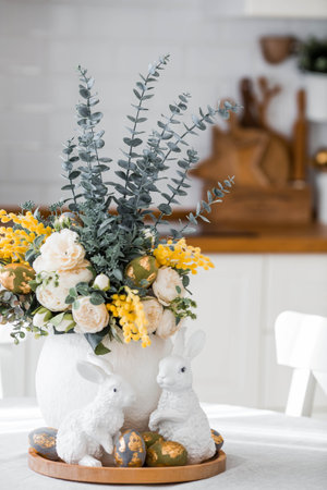A spring bouquet of roses, mimosa and eucalyptus, Easter bunnies and eggs with a golden pattern on the table. A white kitchen is depicted in the background. Beautiful Easter card.の写真素材