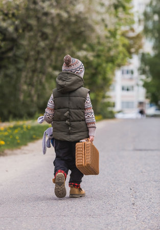 Childhood. A little boy on the street with a suitcase and a stuffed rabbit goes on a journey. Beautiful portrait of a child playing outside.の写真素材