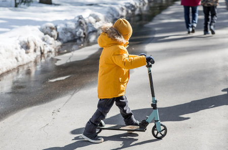 A little boy in bright yellow clothes rides a scooter. A happy child walks outside in early spring.の写真素材