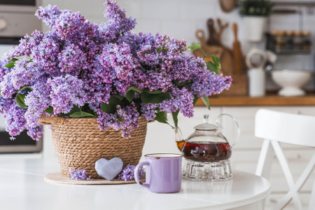 A large purple bouquet of lilacs in a wicker basket, a teapot and a cup of tea on the table. In the background is the interior of a modern white kitchen. The concept of home comfort.の写真素材
