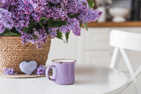 A large purple bouquet of lilacs in a wicker basket, a teapot and a cup of tea on the table. In the background is the interior of a modern white kitchen. The concept of home comfort.の写真素材