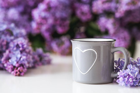 A gray cup of tea with a heart or coffee on the table. In the background is a large purple bouquet of lilacs. The concept of home comfort and drinks.の写真素材