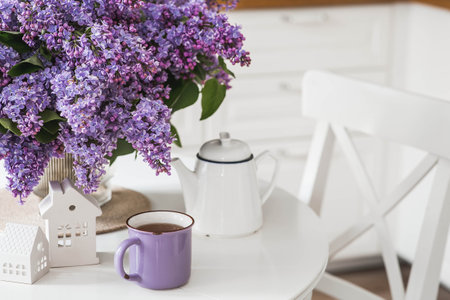 A large purple bouquet of lilacs, white Scandinavian houses, a ceramic teapot and a cup on the table. In the background is the interior of a modern white kitchen. The concept of home comfort.の写真素材