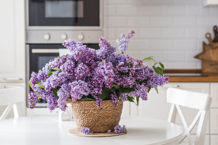 A large purple bouquet of lilacs on the table in a wicker basket. In the background is the interior of a modern white kitchen. The concept of home comfort and celebration.の写真素材