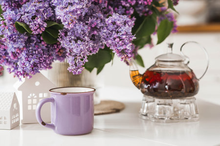 A large purple bouquet of lilacs, white Scandinavian houses, a ceramic teapot and a cup on the table. In the background is the interior of a modern white kitchen. The concept of home comfort.の写真素材