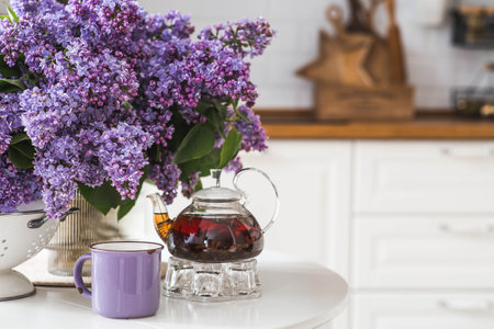 A large purple bouquet of lilacs, a teapot and a cup of tea on the table. In the background is the interior of a modern white kitchen. The concept of home comfort.の写真素材