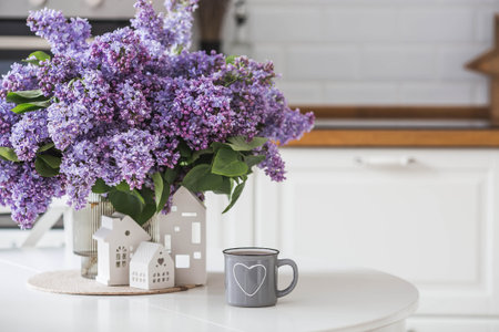A large purple bouquet of lilacs, white Scandinavian houses and a cup of tea on the table. In the background is the interior of a modern white kitchen. The concept of home comfort.の写真素材