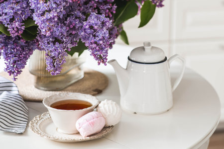 A large purple bouquet of lilacs, a ceramic teapot, a cup and a delicious marshmallow on the table. In the background is the interior of a modern white kitchen. The concept of home comfort.の写真素材