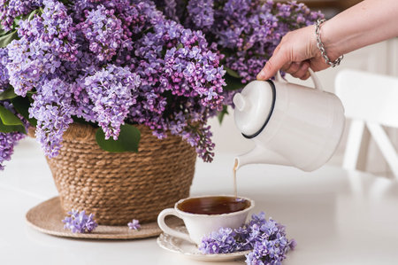 A purple bouquet of lilacs in a wicker basket and a woman's hand pours tea into a mug from a teapot. In the background is the interior of a modern white kitchen. The concept of home comfort.の写真素材