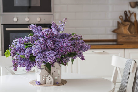 A large purple bouquet of lilacs and white Scandinavian houses on the table. In the background is the interior of a modern white kitchen. The concept of home comfort.の写真素材