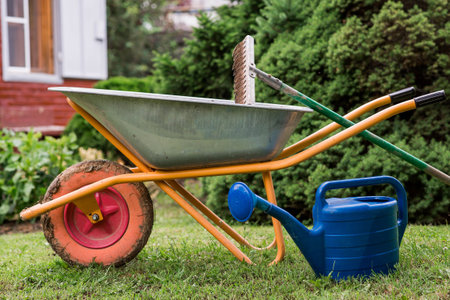 A garden wheelbarrow, rake, and watering can in the backyard of a house. The concept of gardening and country life. Preparing the garden for autumn and winter. Closing the summer season.の写真素材