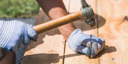 Close-up of a nail being driven into a board with a hammer. A carpenter wearing cloth protective...